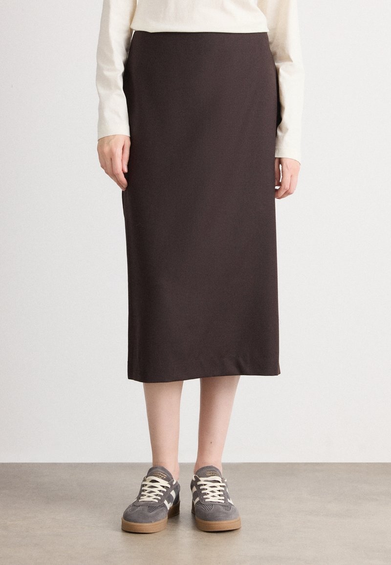 Personne portant une jupe midi marron foncé, un haut à manches longues crème et des baskets en daim gris avec des lacets blancs, debout sur un sol lisse.