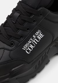 Zapatilla negra con parte superior de cuero suave, suela texturizada, logo prominente "VERSACE JEANS COUTURE" en blanco y cordones redondos.