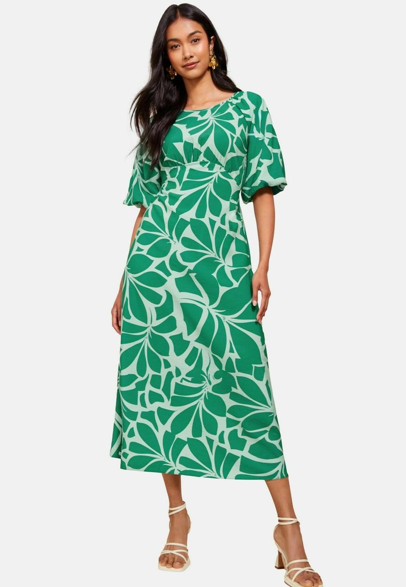 Rochie maxi de culoare verde, cu un imprimeu îndrăzneț de frunze, mâneci scurte bufante și un corset ajustat. Asortată cu sandale cu toc de culoare bej. Textură de țesătură netedă.