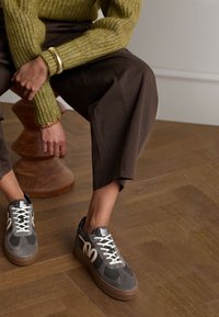 Gray sneakers with white accents and black mesh panels, featuring thick rubber soles and a rounded design. Textured brown flooring and styled clothing.