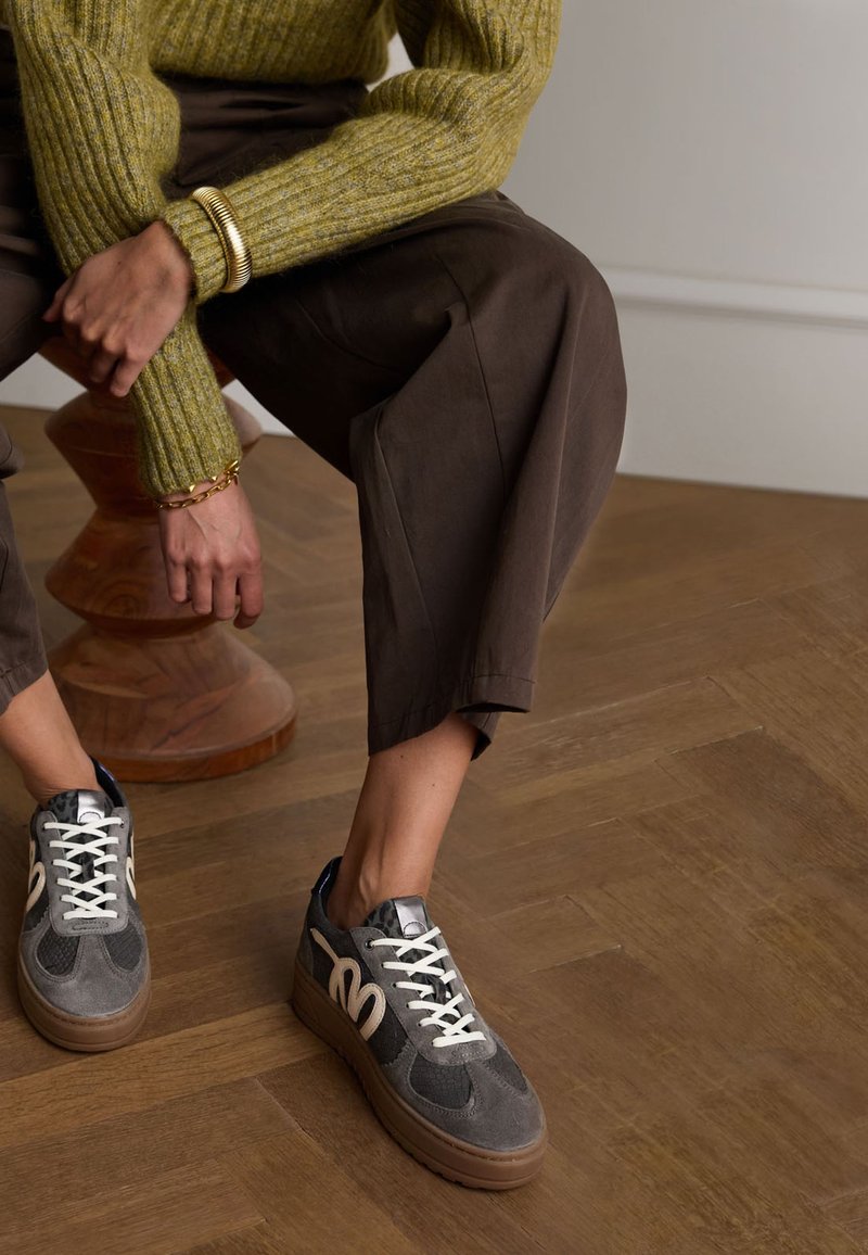 Gray sneakers with white accents and black mesh panels, featuring thick rubber soles and a rounded design. Textured brown flooring and styled clothing.