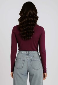 Top bordeaux a maniche lunghe abbinato a jeans azzurri a vita alta, caratterizzati da una vestibilità rilassata e tasche posteriori. Capelli mossi e sciolti.