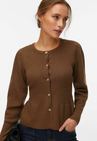 Femme portant un cardigan marron à manches longues boutonné avec des boutons dorés, un jean bleu foncé, tenant un sac noir, regardant sur le côté.