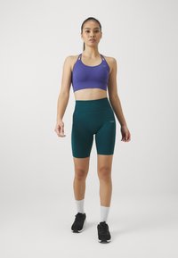 Fioletowy stanik sportowy i turkusowe wysokie spodenki rowerowe. Cechy obejmują elastyczny materiał, dopasowany krój oraz boczną kieszeń w spodenkach.