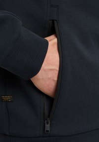 Dunkelblaues Sweatshirt mit einer Reißverschlusstasche, gerippten Bündchen und einer weichen Stofftextur. Reißverschlussdetail an der Tasche sichtbar.