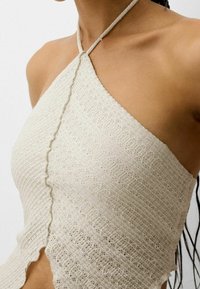 Jasno beżowy dzianinowy top na halterze z falbankami i teksturowym wzorem, z cienkimi ramiączkami wokół szyi.
