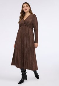 Bronzefärgad klänning med lång ärm, V-ringning och vridet knutdetalj vid midjan. Flytande, plisserad kjol med slät textur. Bärs med svarta knähöga stövlar.