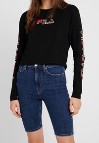 Černá dlouhá rukávová košile s logem v leopardím potisku a červenými akcenty, spárovaná s modrými denimovými bermudami, které mají přiléhavý střih a středně vysoký pas.