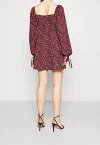 Punainen kukallinen mekko, jossa on pitkät, pöyhöiset hihat ja neliönmuotoinen pääntie. Mekossa on kireä vyötärö ja liehuva hame. Yhdistetty mustiin korkeakorkoisiin sandaaleihin.