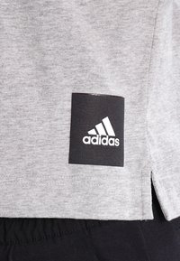 Szara bawełniana koszulka z czarną łatką z białym logo "adidas" na dole; szczeliny po bokach i miękka tekstura.