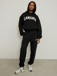 Juodas džemperis su gobtuvu ir atitinkamos spalvos sportinės kelnės, kuriose yra baltas užrašas "LAMANIA". Pagaminta iš minkštos medžiagos, su laisvu pjūviu, derinama su sportiniais batais.