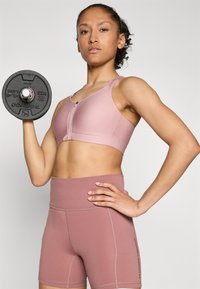 Sujetador deportivo rosa claro con cremallera frontal, combinado con pantalones cortos de cintura alta a juego. La modelo sostiene un peso negro en una mano.