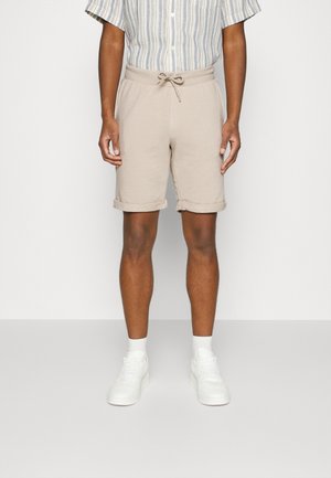 Homme portant un short beige à cordon, des baskets blanches et une chemise rayée à manches courtes boutonnée, debout devant un fond uni.