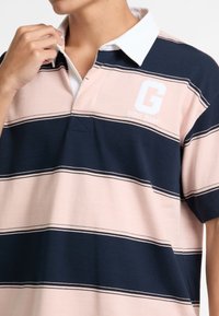 Polo tričko v ružových a námorníckych pruhách s bielym golierom. Má malé biele logo "G" a vyšívaný nápis "GUESS JEANS" na hrudi.