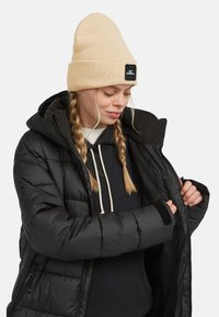 Zwarte pufferjack met capuchon, ritssluitingzakken en duimgaten in de mouwen. Draag het over een marineblauwe hoodie, afgewerkt met een lichtbeige muts.