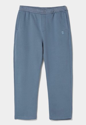 Niebieskie spodnie dresowe z elastycznym pasem, bocznymi kieszeniami i małym wyszywanym logo na górnej lewej udzie, pokazane na płasko na białym tle.