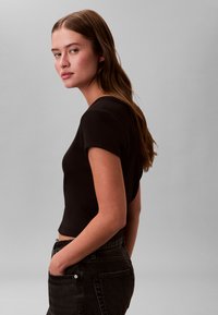 Noor pruunide juustega naine, kes kannab musta lühikeste varrukatega pluusi ja musti teksaseid, seisab küljelt, käed taskutes, halli tausta ees.