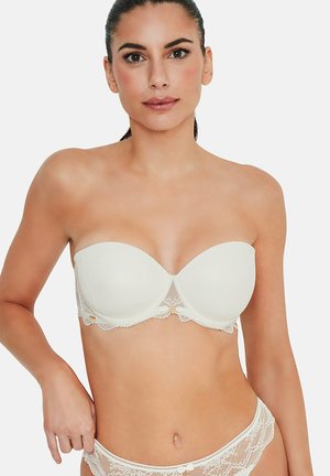 Femme portant un soutien-gorge en dentelle blanc sans bretelles et une culotte assortie en dentelle, posant avec la main sur la hanche devant un fond uni.