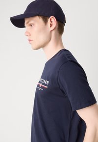 Navy T-shirt met een ronde hals, voorzien van een wit en rood gedrukte logo en tekst. Gecombineerd met een bijpassende marineblauwe pet.