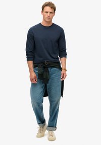 Camisa de manga larga azul marino, jeans de corte relajado en un tono azul claro y zapatillas de color crema. Una camisa verde oscura atada a la cintura añade contraste.