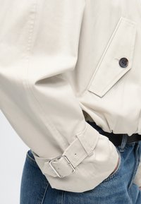 Μανίκι μπεζ σακακιού με τσέπη με καπάκι και κουμπί, ρυθμιζόμενο μανσέτα με πόρπη, συνδυασμένο με μπλε τζιν και μαύρη ζώνη.