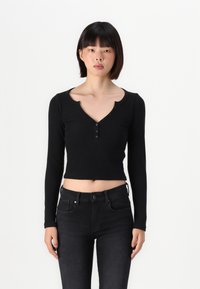 Černý dlouhý rukáv cropped top s výstřihem do V a zapínáním na knoflíky; vyrobeno z žebrovaného materiálu; nošeno s tmavými denimi.