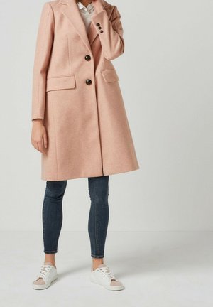 Vrouw draagt een knie-omvattende wollen jas in blushroze, donkere strakke jeans, witte sneakers en een wit overhemd met kraag onder de jas.