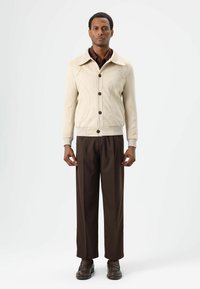 Lichtbeige jas met contrasterende naden, vijf knopen en een omslagkraag, gecombineerd met donkerbruine geplooide broek en bruine schoenen.