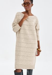 Robe en tricot beige avec des rayures horizontales et des détails en œillets. Présente un large col bateau et des poignets côtelés. Associée à des bottes noires montantes.