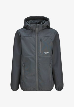 Junge mit lockigem Haar, der eine graue Kapuzenjacke mit Reißverschluss, weite blaue Jeans und braune Schuhe trägt und vor einem einfarbigen Hintergrund steht.