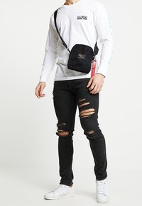 Czarna torebka na ramię z przodu z logo, czerwona zawieszka na suwaku; noszona z białą koszulą z długim rękawem, postrzępionymi czarnymi dżinsami i białymi trampkami.