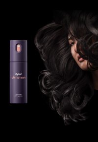 Spray per styling multiuso Dyson in bottiglia viola scuro con accenti rosa; presenta una forma cilindrica e un ugello spray. Capelli neri fluenti sullo sfondo.