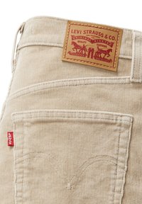 Pantaloni in corduroy beige con cuciture a contrasto, dotati di tasche posteriori e un'etichetta in pelle Levi Strauss con dettagli in rilievo. Tag rosso sul lato.