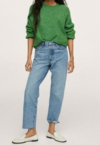 Grüner Strickpullover, hellblaue Jeans mit hoher Taille und weiße Slip-On-Schuhe. Mit hochgekrempelten Ärmeln und einem kurzen Schnitt an den Knöcheln.