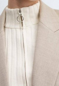 Blazer beige sartorial con un top bianco a zip con righe e un anello di chiusura argentato. Tessuto testurizzato con righe verticali lungo la zip.