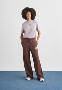 Ανοιχτό μωβ κοντομάνικη μπλούζα με στάμπα «Nike Shox», συνδυασμένη με καφέ, φαρδιά παντελόνια φούτερ με λογότυπο. Το σύνολο ολοκληρώνεται με λευκά αθλητικά παπούτσια.