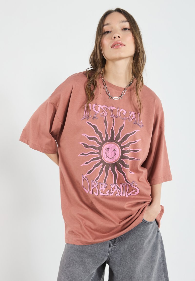 Overdimensioneret lerfarvet t-shirt lavet af bomuld, med et stort pink solgrafik og teksten "Mystical Dreams" i en legende skrifttype.