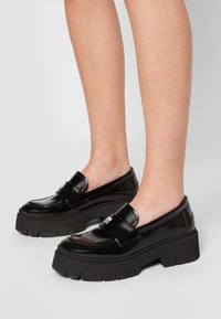 Mocassins en cuir verni noir avec des semelles compensées noires épaisses, featuring un bout arrondi et une sangle décorative. Détails de couture texturée visibles.