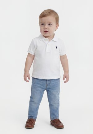 Bambino con capelli chiari che indossa una polo bianca, jeans azzurri e scarpe di pelle marrone, in piedi su uno sfondo bianco.