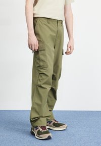 Olive green cargo pants with multiple side pockets, straight-leg cut, and textured fabric, paired with multi-color athletic shoes.