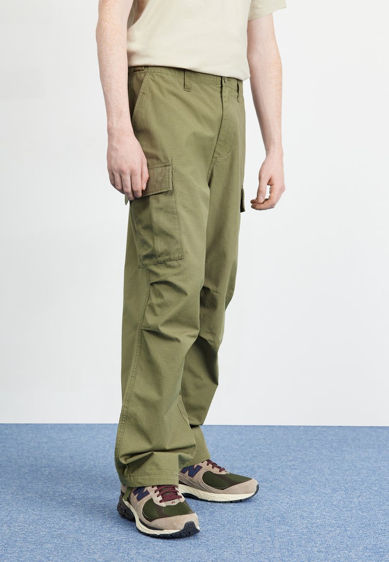 Olive green cargo pants with multiple side pockets, straight-leg cut, and textured fabric, paired with multi-color athletic shoes.