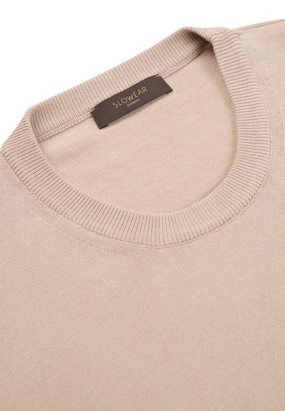 Suéter beige de punto con cuello redondo, tela texturizada y una etiqueta marrón oscuro que dice "SLOWEAR" colocada en el cuello.