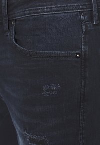 Tummansiniset farkut, joissa on kulutettuja kohtia, hailakkaa kulutusta, pyöristetyillä reunoilla varustettu etutasku ja pieni hopeinen nappi.