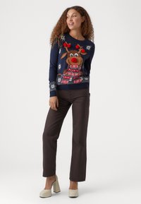 Pulover de Crăciun bleumarin cu un design de ren, decorat cu pompoane roșii, fulgi de zăpadă și modele colorate la tiv. Asortat cu pantaloni maro.