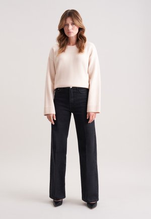 Frau steht und trägt einen cremefarbenen Pullover, hochtaillierte schwarze weite Jeans und schwarze spitze Schuhe vor einem einfarbigen hellen Hintergrund.