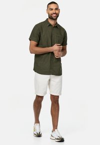 Kurzärmliges olivgrünes Hemd mit Knopfleiste, kombiniert mit weißen hochgekrempelten Shorts und weißen Sneakers mit grauen und gelben Akzenten.