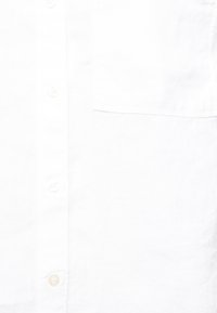 Biała lniana koszula z guzikiem, z kieszonką, o miękkiej fakturze i subtelnym połysku. Szczegóły guzików są w jasnym kolorze i lekko przezroczyste.