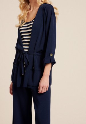 Woman wearing navy blue belted jacket with roll-tab sleeves over black and white striped top and matching navy wide-leg pants.