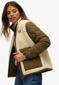 Una donna che sistema una giacca in pile verde e crema con maniche e tasche frontali trapuntate, mentre indossa jeans blu su uno sfondo bianco.