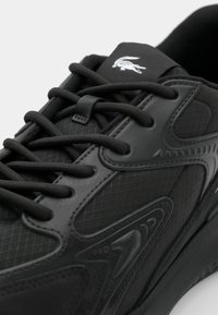 Gros plan d'une basket noire avec une surface texturée, des lacets noirs et un logo crocodile blanc sur la languette.